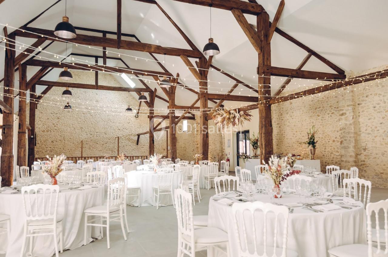Salle de réception lumineuse avec murs en pierre, poutres apparentes, tables rondes dressées et guirlandes lumineuses.