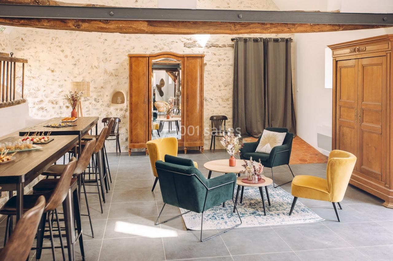 Salle lumineuse avec murs en pierre, mobilier moderne et ancien, table haute, fauteuils colorés et décorations florales.