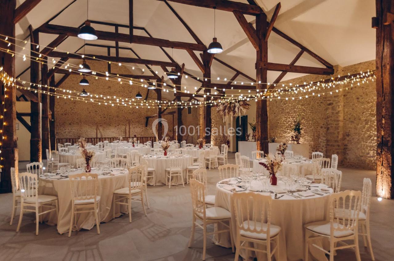 Salle de réception avec tables rondes dressées, chaises blanches et guirlandes lumineuses suspendues sous une charpente en…