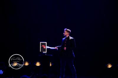 Miniature Edi Cappera #5 Un magicien sur scène tenant une baguette devant un accessoire éclairé dans un décor sombre.