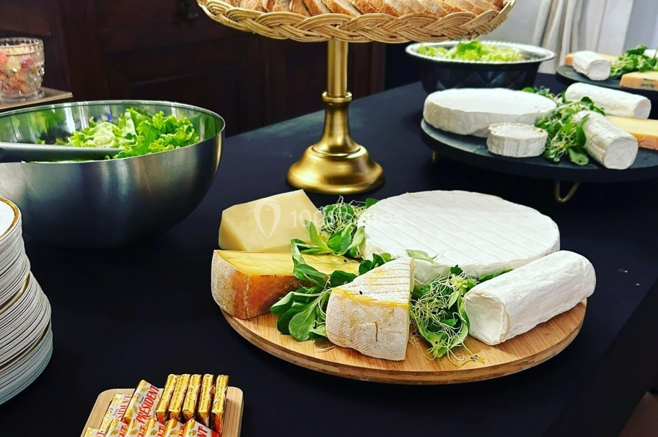 Plateau de fromages variés avec herbes fraîches, pain en corbeille et salade en arrière-plan sur une table noire.