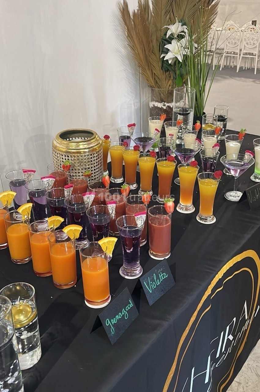 Table garnie de cocktails colorés avec décorations fruitées, étiquettes de saveurs et éléments décoratifs en arrière-plan.