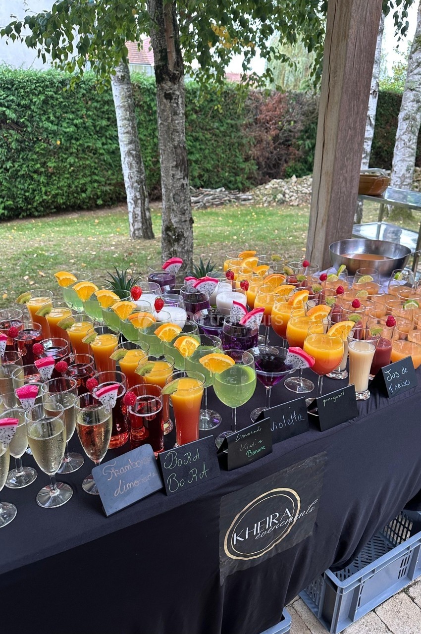 Table garnie de verres de cocktails colorés et de jus, disposés sur une nappe noire en extérieur près d'un jardin.