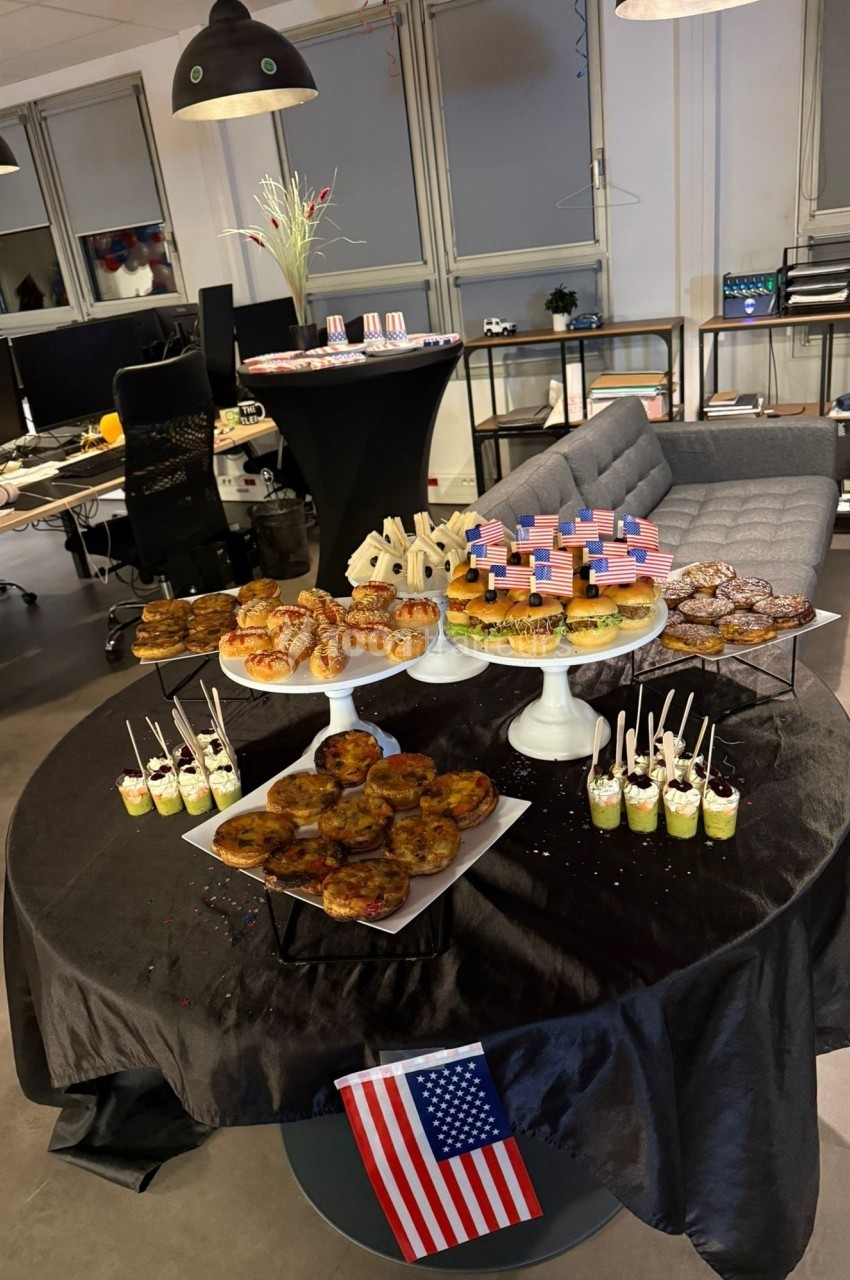 Table garnie de desserts variés, décorés de drapeaux américains, dans un espace de bureau moderne.