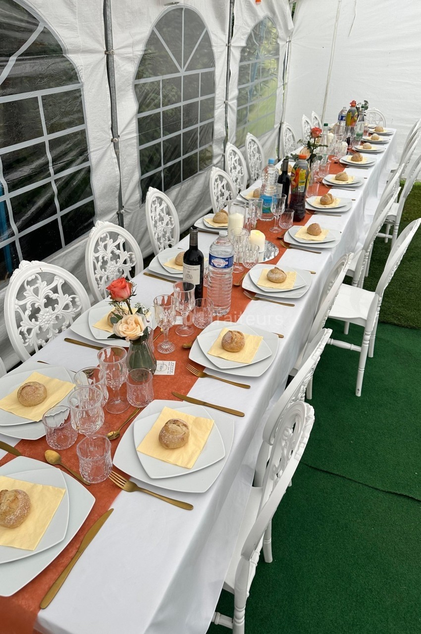 Table dressée sous une tente avec nappes blanches, vaisselle, verres, pains individuels et décorations florales.