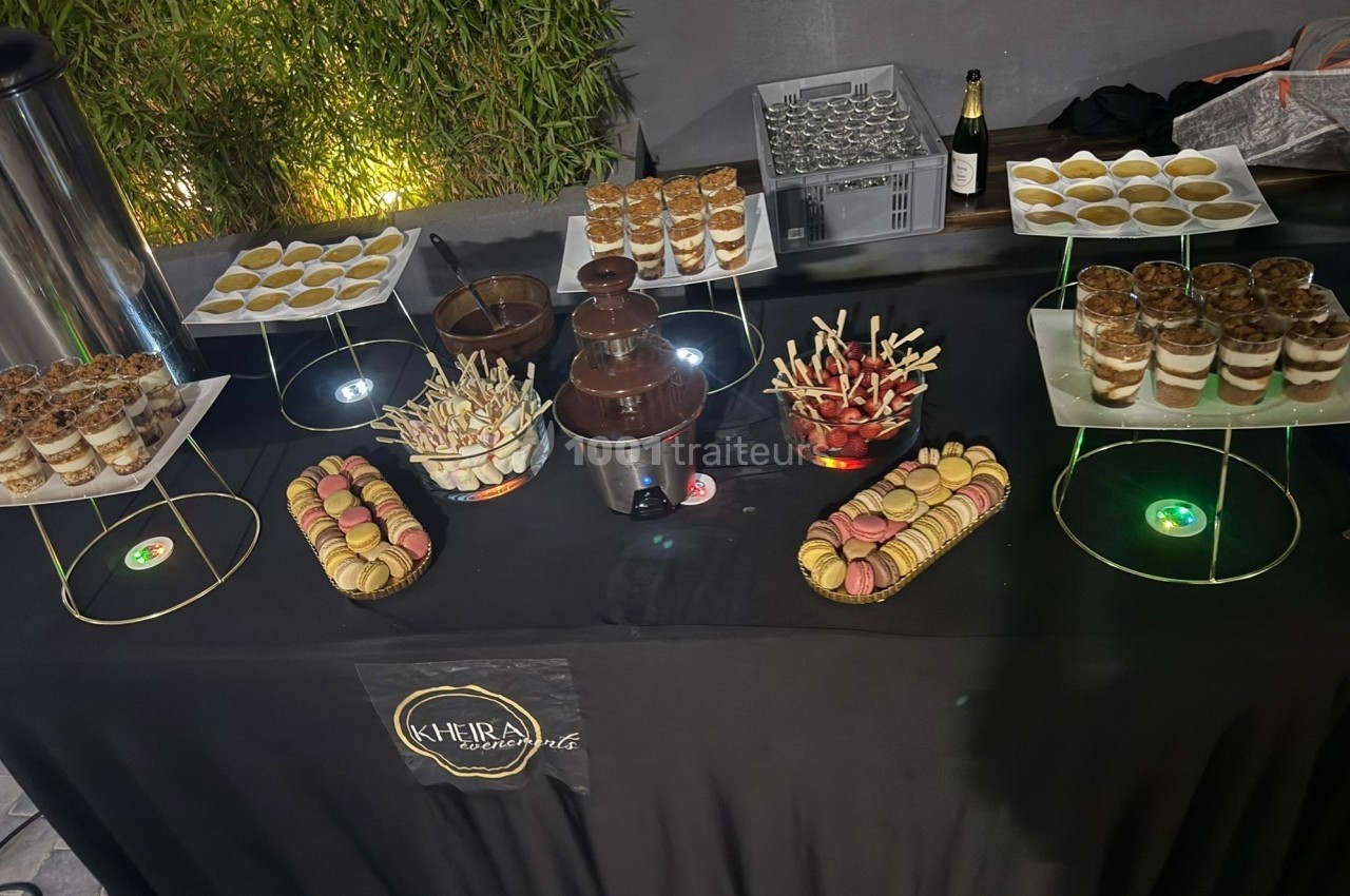 Table de desserts avec macarons, verrines, brochettes de fruits, fontaine de chocolat et boissons en arrière-plan.