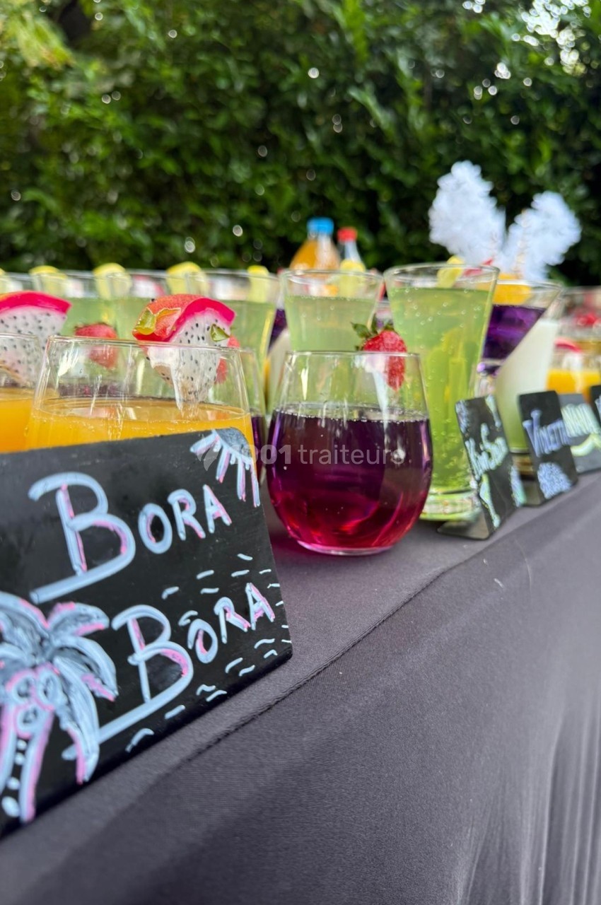 Verres de cocktails colorés alignés sur une table avec des ardoises décorées, en extérieur près de végétation.