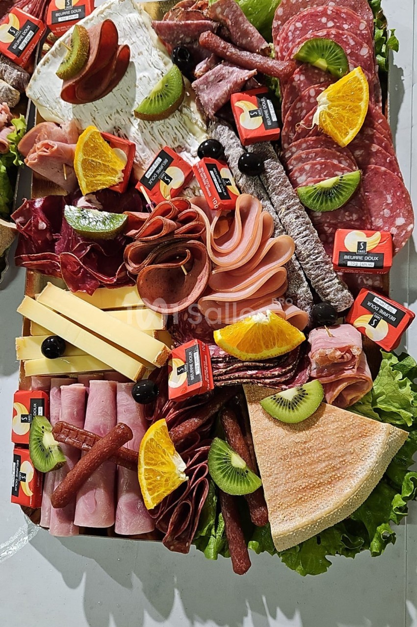 Plateau de charcuterie et fromages variés avec fruits frais, olives et tranches de pain sur un lit de salade verte.