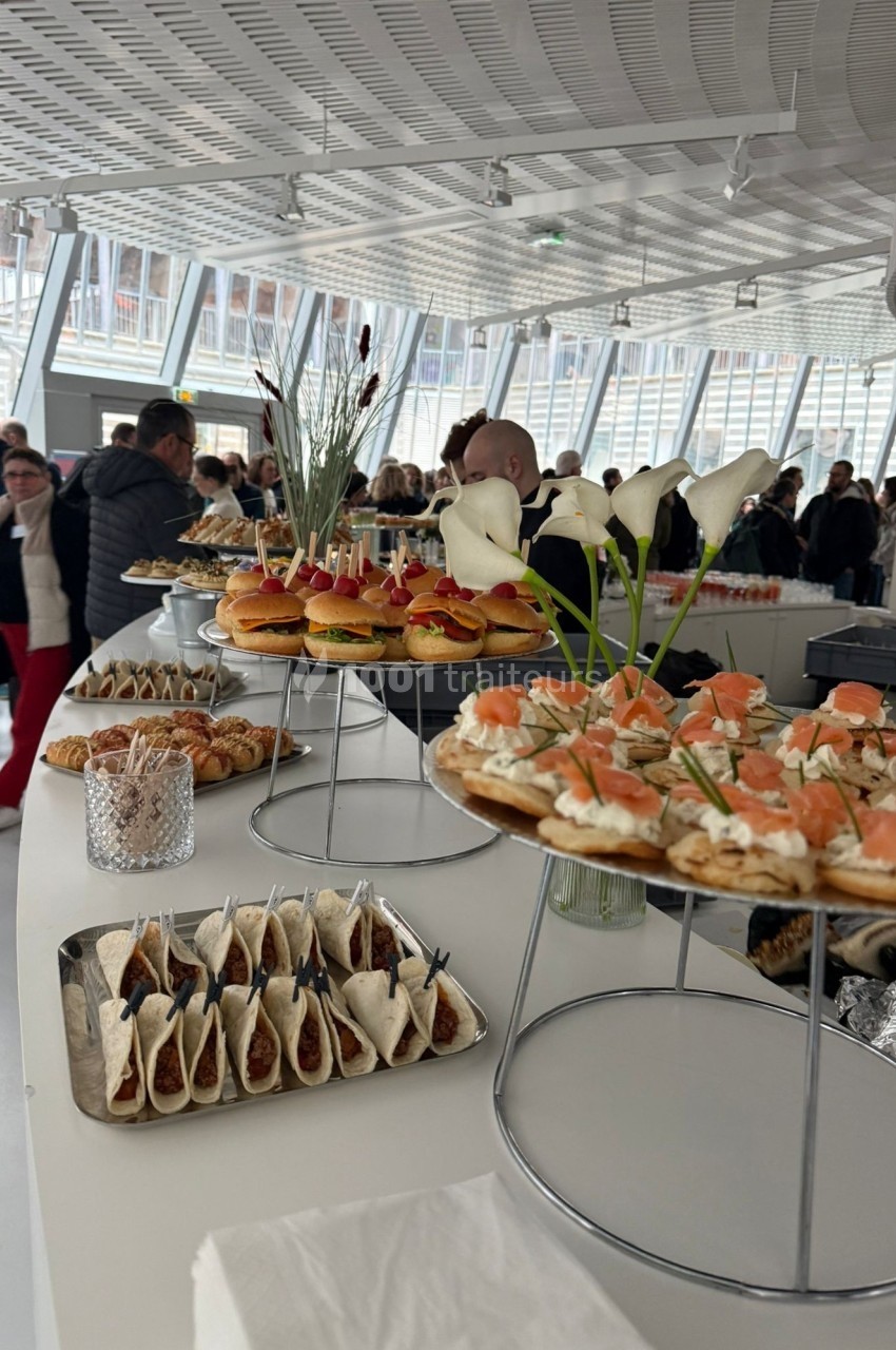 Buffet avec plateaux de canapés, tacos et viennoiseries, dans une salle lumineuse avec des invités en arrière-plan.