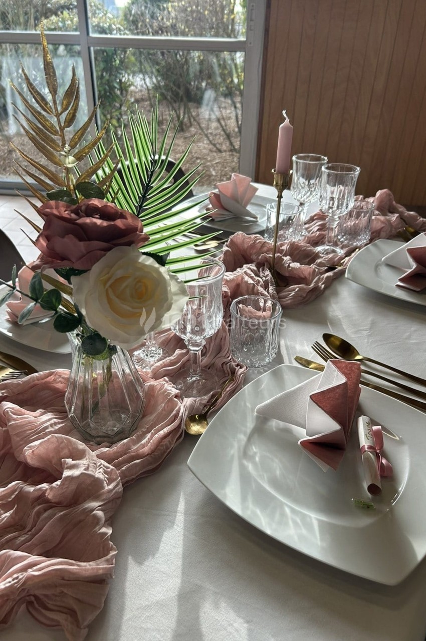 Table décorée avec des assiettes blanches, serviettes roses pliées, verres, bougies et un bouquet de fleurs dans un vase.