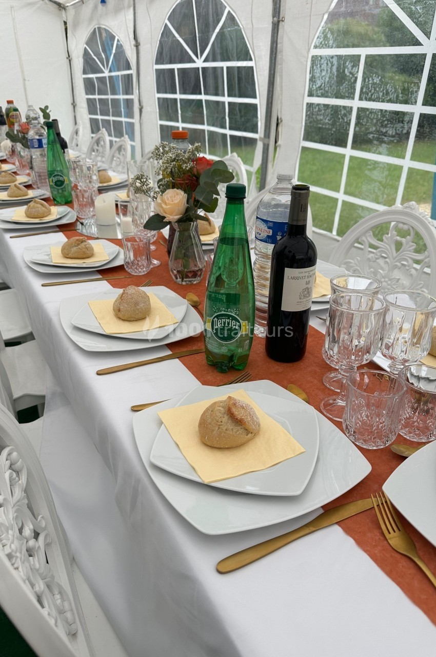 Table dressée sous une tente avec assiettes, verres, pains, bouteilles d'eau et de vin, décorée de fleurs.