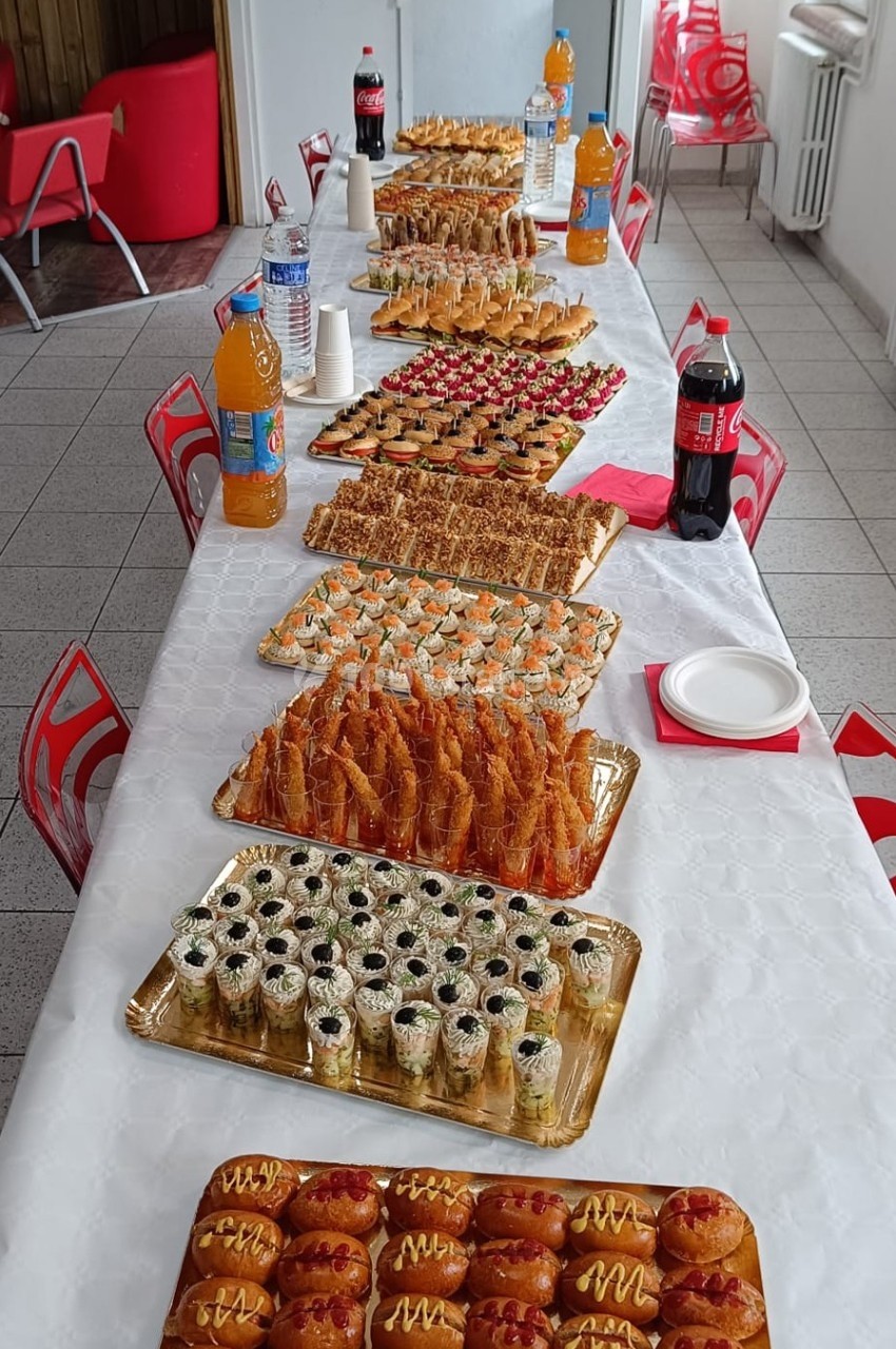 Table garnie de plateaux de pâtisseries, sushis et amuse-bouches, accompagnés de boissons, dans une salle lumineuse.