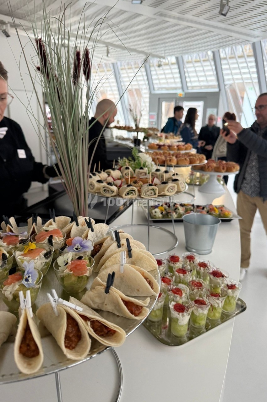 Buffet varié avec wraps, verrines et amuse-bouches, présenté dans une salle lumineuse avec des convives en arrière-plan.