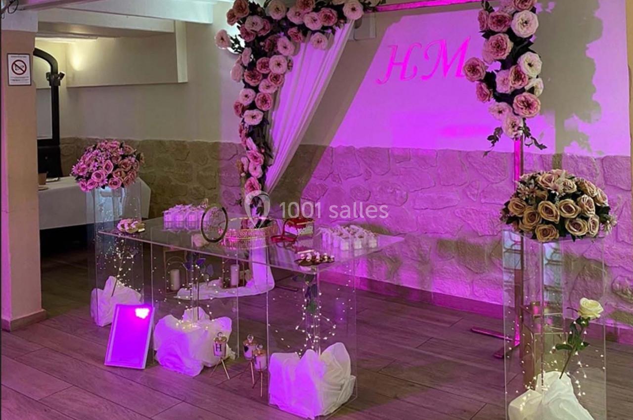 Décoration de table de fête avec fleurs roses, guirlandes lumineuses et rideaux blancs dans une salle éclairée en rose.