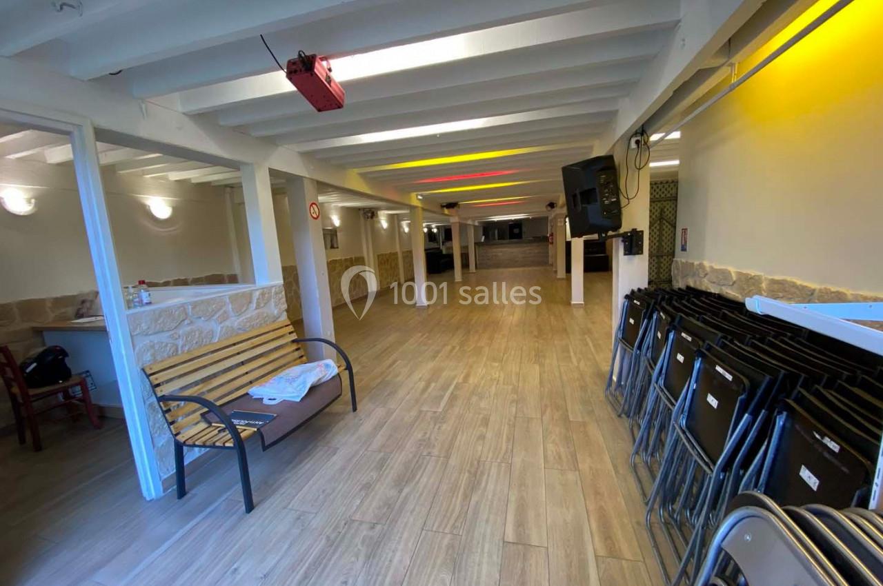 Salle vide avec sol en bois, banc, chaises empilées à droite, murs clairs et éclairage jaune au plafond.