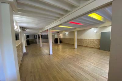 Salle spacieuse avec sol en parquet, murs en pierre et poutres blanches apparentes, éclairée par des lumières modernes.