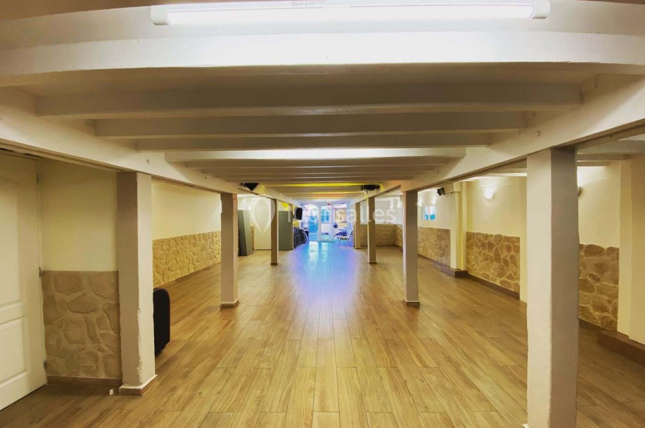 Salle vide avec sol en parquet, murs clairs partiellement recouverts de pierre, et poutres blanches au plafond.