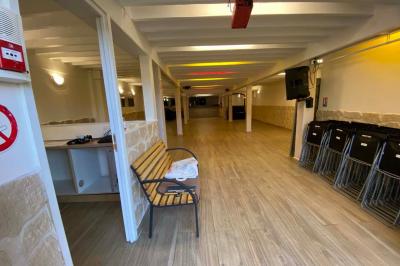 Salle spacieuse avec sol en parquet, murs en pierre et poutres blanches apparentes, éclairée par des lumières modernes.