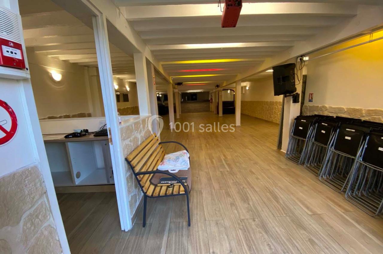 Salle vide avec sol en bois, murs clairs, rangées de chaises empilées et un banc en bois près de l'entrée.