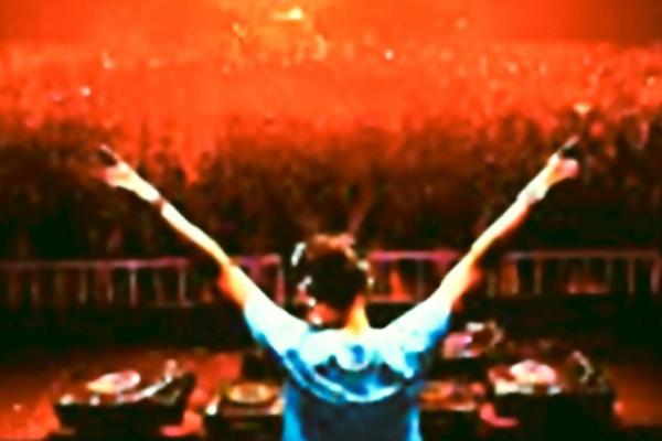 Un DJ de dos lève les bras devant une foule dense dans une ambiance lumineuse rouge.