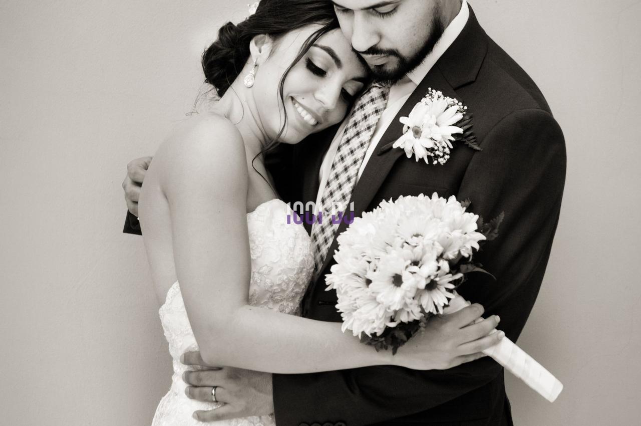 Un couple en tenue de mariage s'enlace tendrement devant un mur clair.