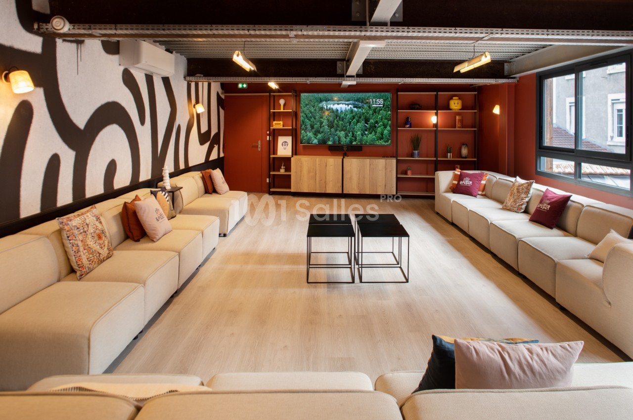 Salle de séjour spacieuse avec canapés beiges, tables basses noires, étagères décorées et télévision allumée.