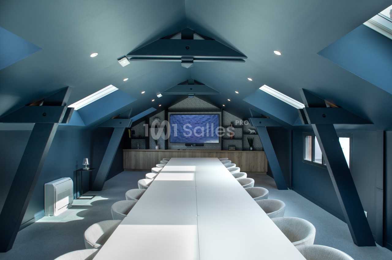 Salle de réunion moderne sous combles, avec une longue table blanche, chaises grises et écran mural au fond.