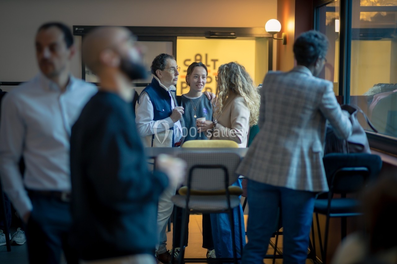 Des personnes discutent debout dans un espace intérieur lumineux, près de tables hautes et d'une baie vitrée.