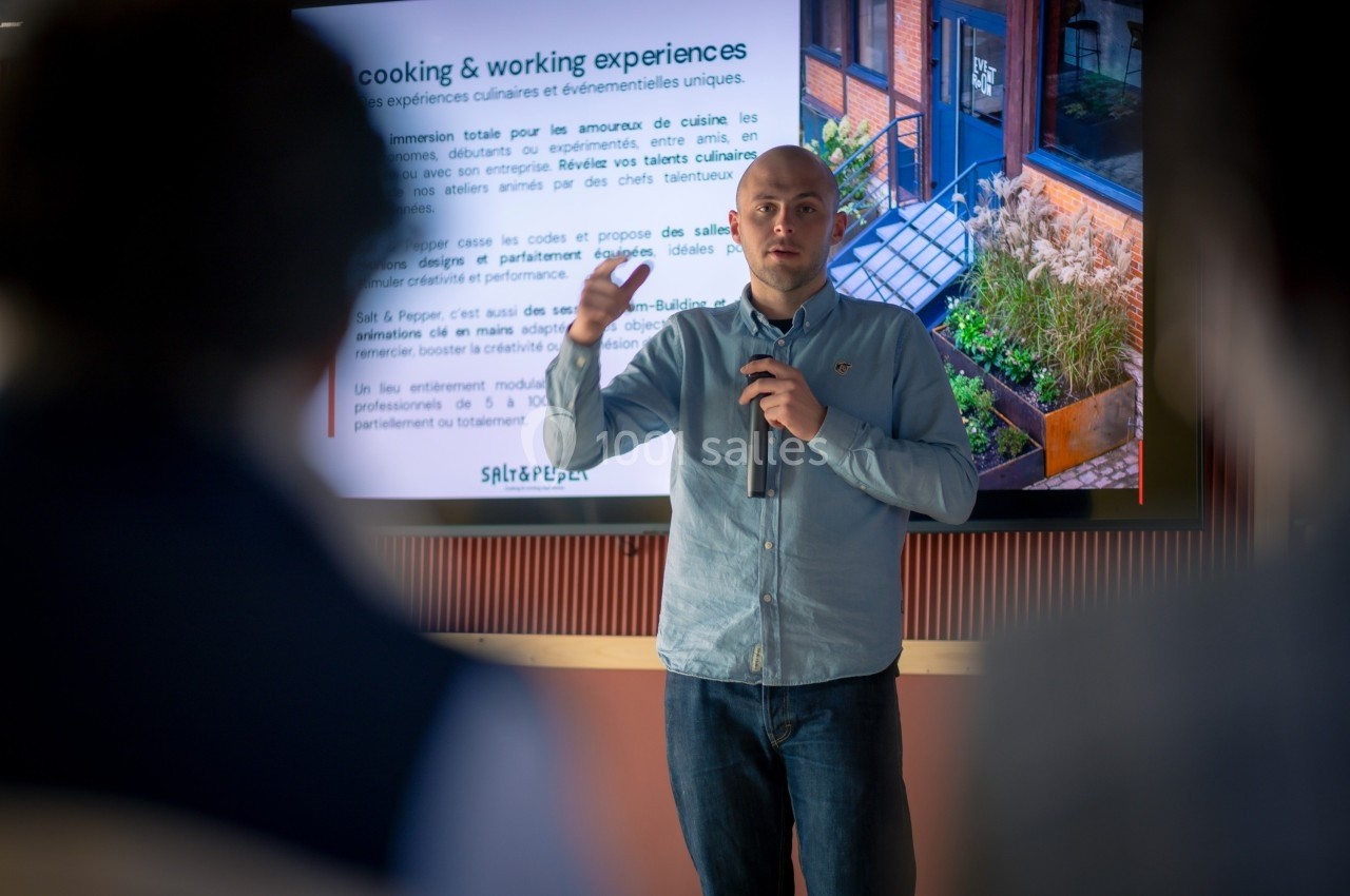 Un homme debout parle dans un micro devant un écran affichant une présentation dans une salle.