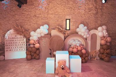 Décor de fête avec ballons beige et blanc, murs en pierre, présentoirs et un ours en peluche au centre.