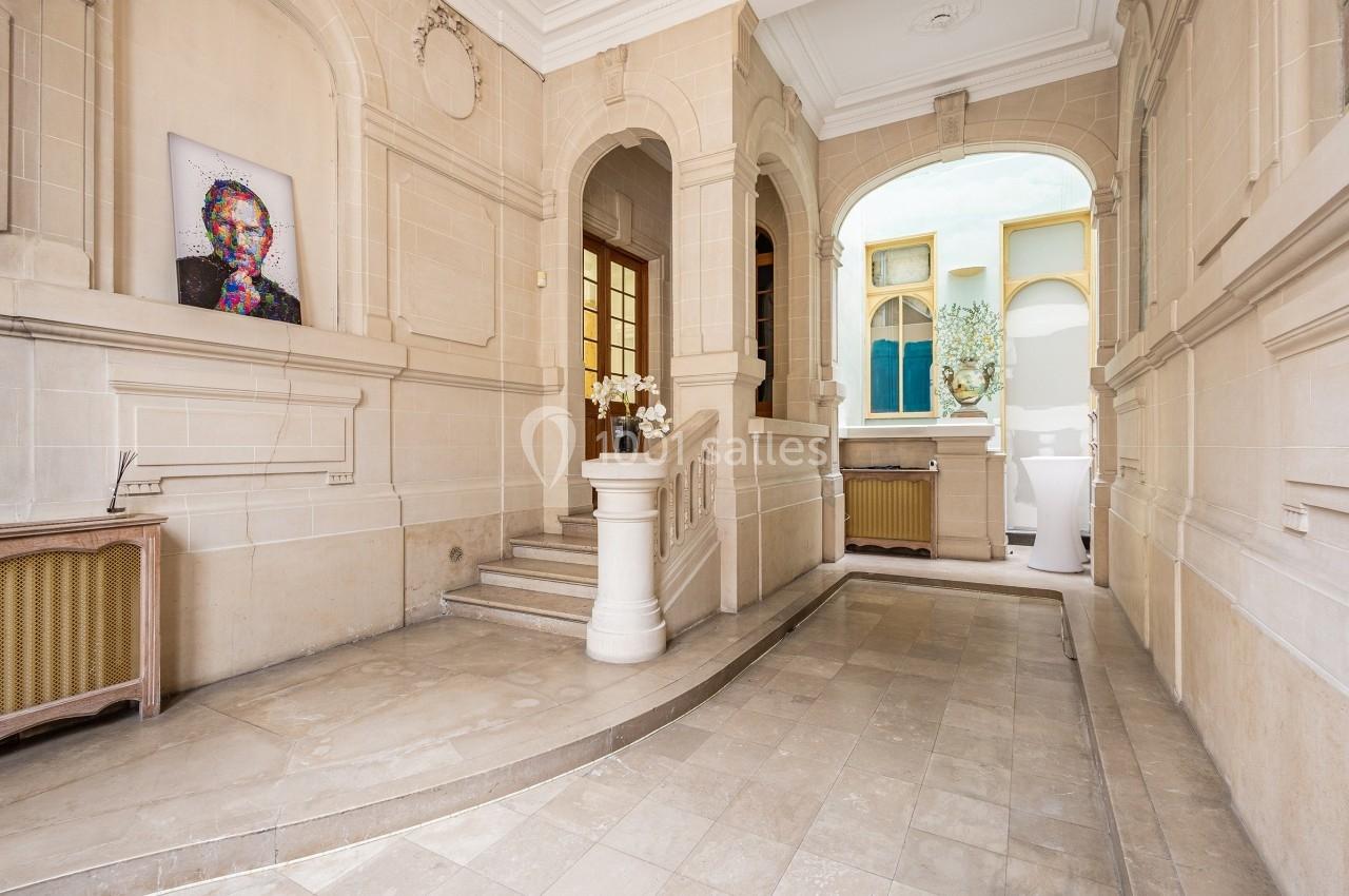 Entrée lumineuse avec murs en pierre, tableau coloré, escalier, arches et vue sur une cour intérieure.