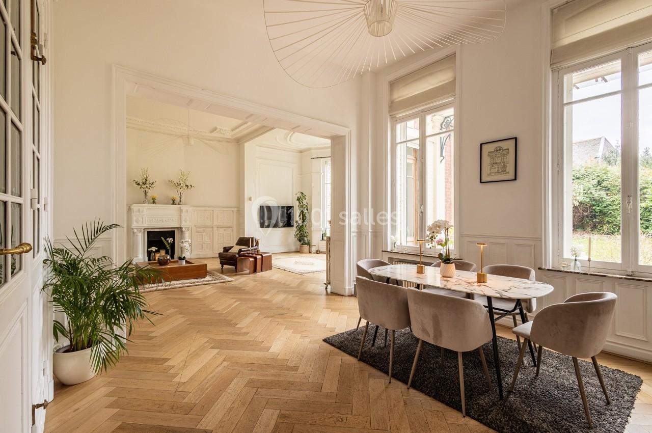Salle à manger lumineuse avec table en marbre, chaises grises, parquet en bois clair et vue sur un salon avec cheminée.