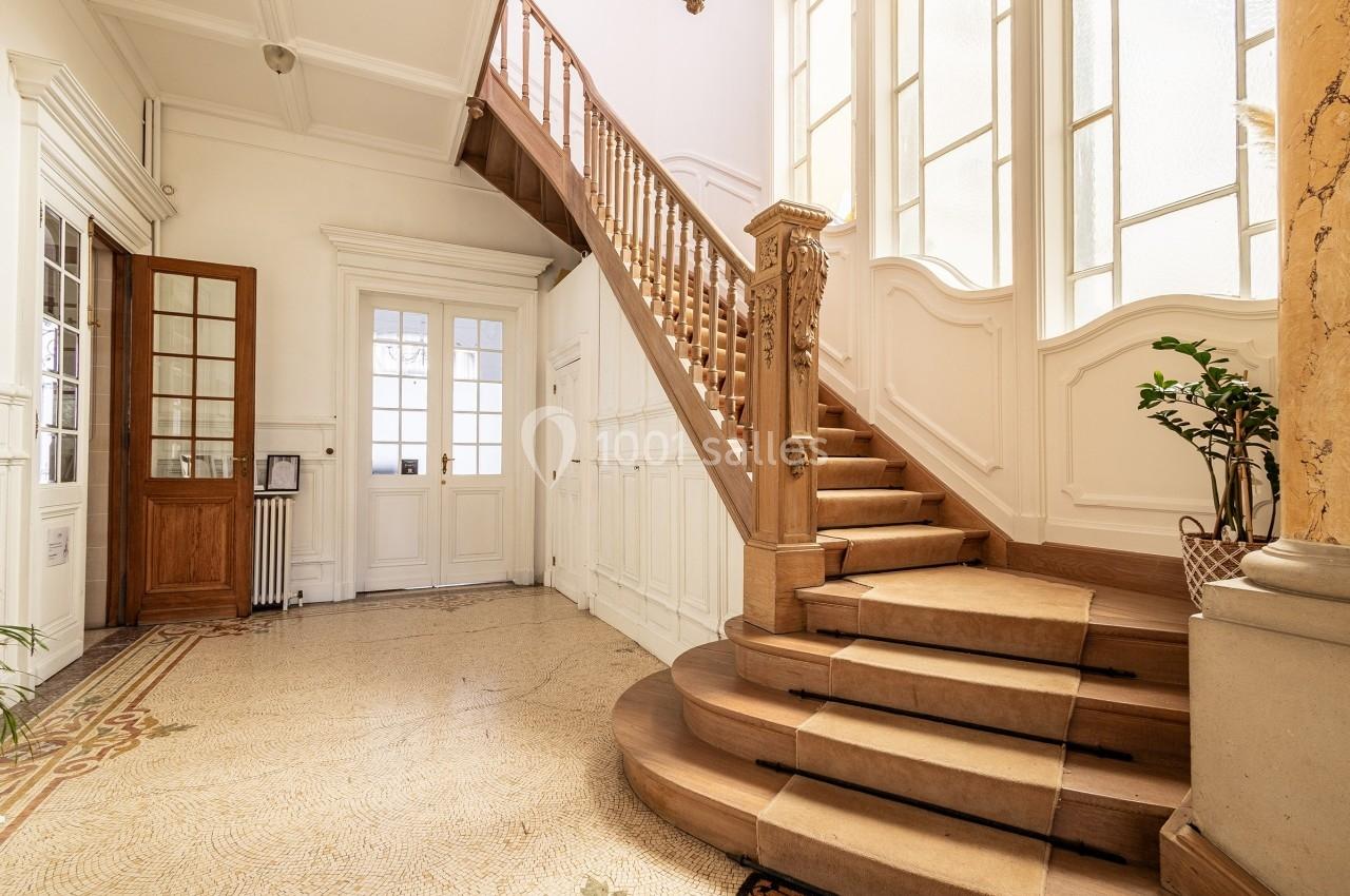 Escalier en bois dans un hall lumineux avec sol en mosaïque, murs blancs et grandes fenêtres laissant entrer la lumière.