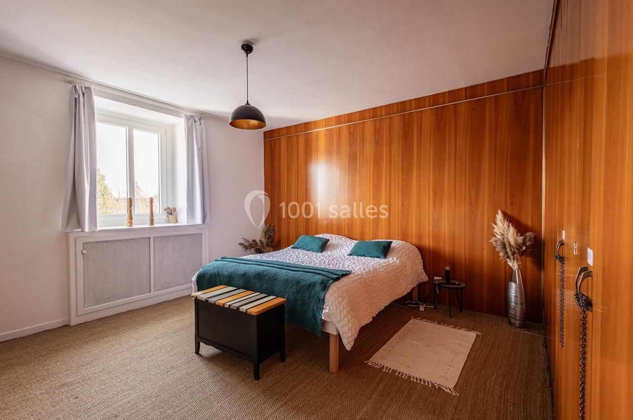 Chambre lumineuse avec lit double, mur en bois, tapis en jonc de mer, fenêtre avec rideaux blancs et décoration minimaliste.