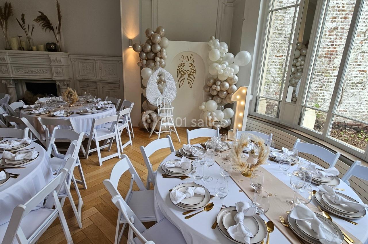 Salle décorée pour un événement avec tables dressées, nappes blanches, ballons et panneau lumineux près d'une fenêtre.