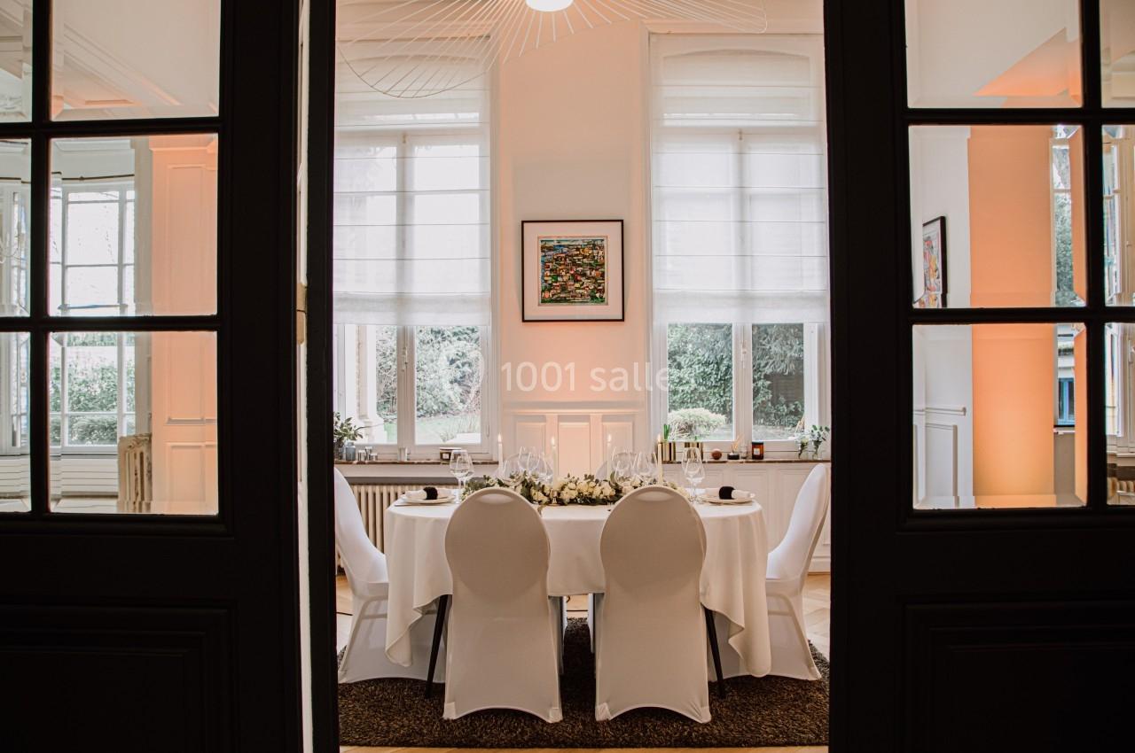 Salle élégante avec table ronde dressée pour un repas, entourée de chaises blanches et éclairée par des fenêtres.