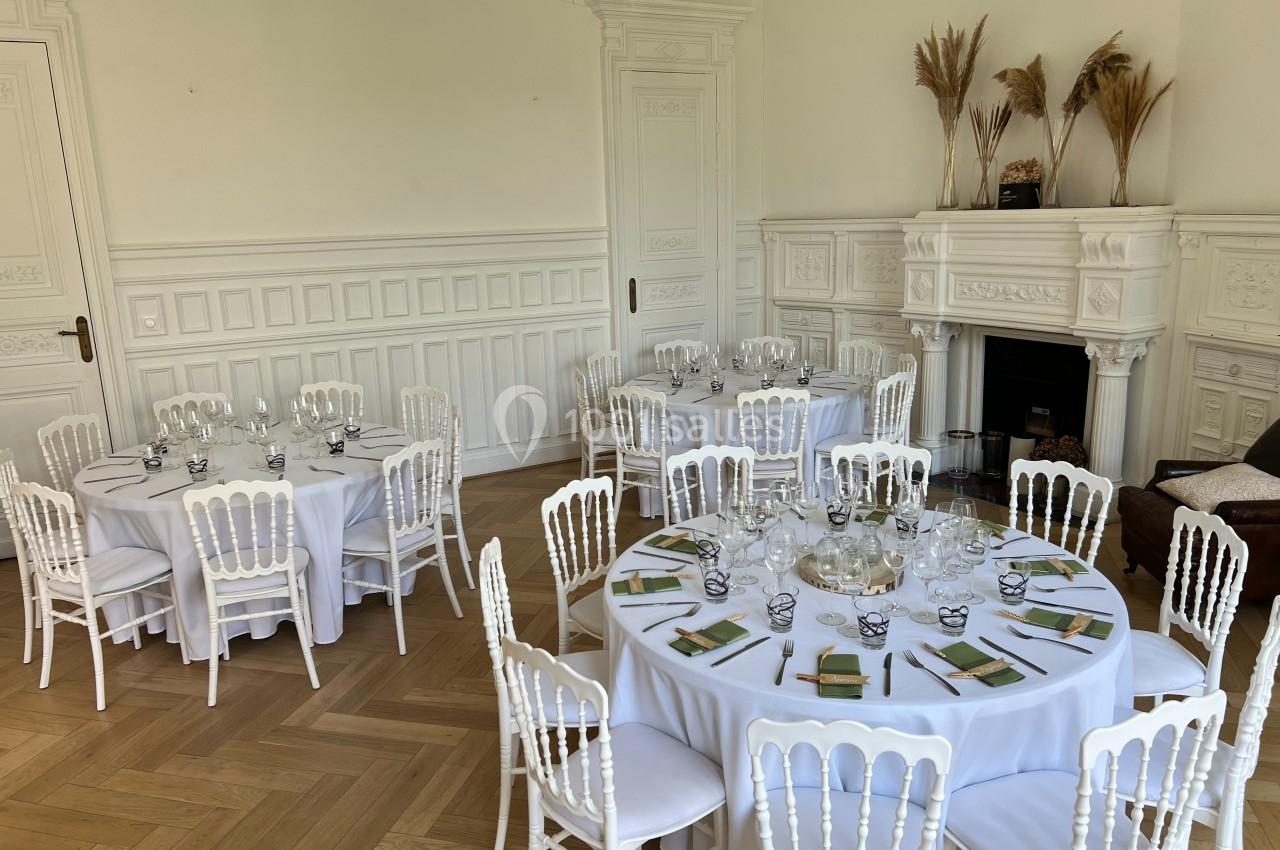 Salle élégante avec tables rondes dressées, chaises blanches, parquet clair et cheminée décorative en arrière-plan.