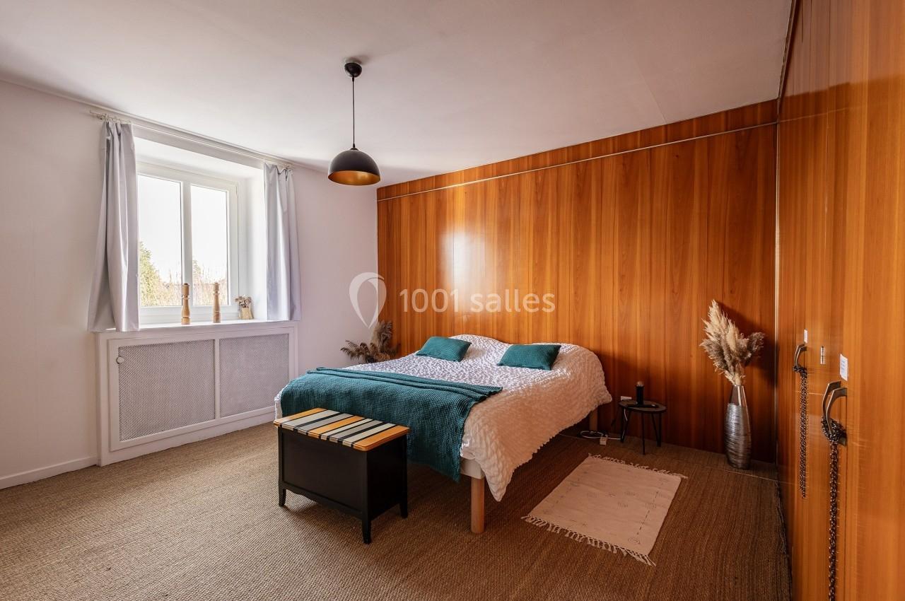 Chambre lumineuse avec lit double, mur en bois, tapis en jute, fenêtre avec rideaux blancs et décoration minimaliste.