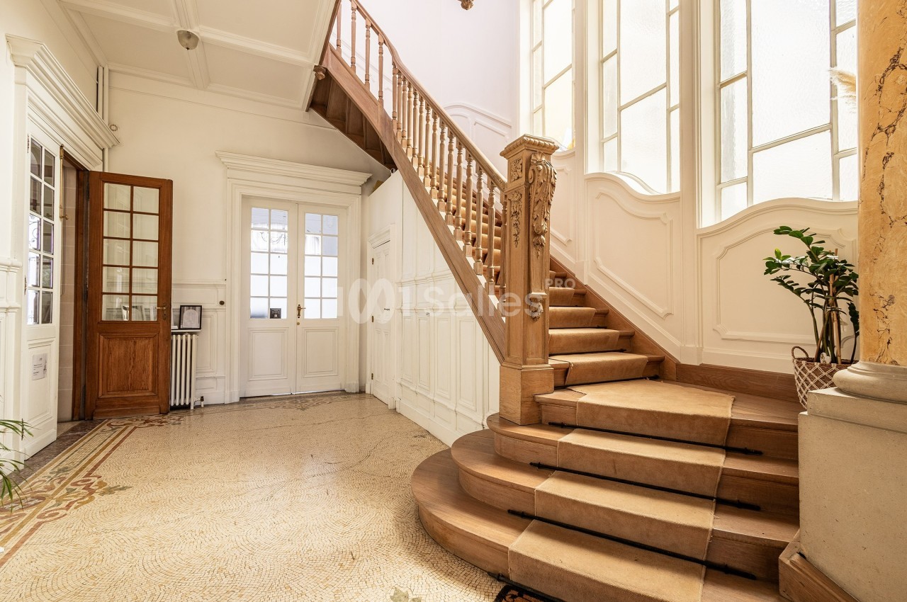 Escalier en bois sculpté dans un hall lumineux avec sol en mosaïque, grandes fenêtres et plantes décoratives.