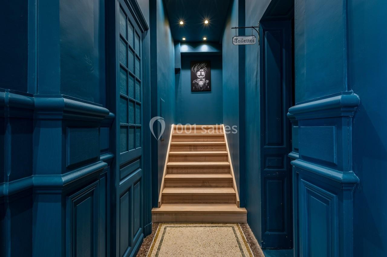 Escalier en bois éclairé menant à un étage, encadré par des murs bleus et un panneau indiquant ’Toilettes’.