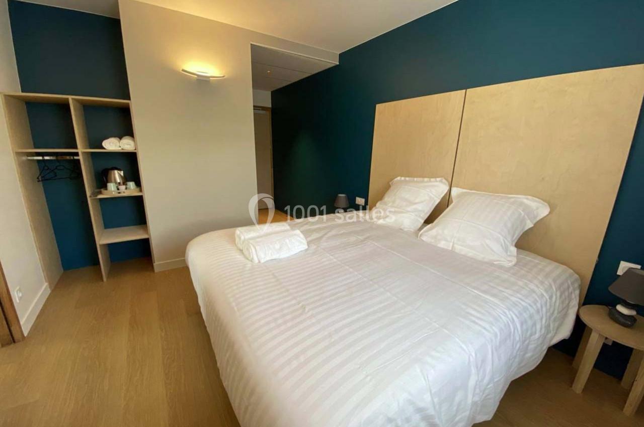 Lit double avec draps blancs dans une chambre moderne, mur bleu, éclairage mural et espace de rangement ouvert.
