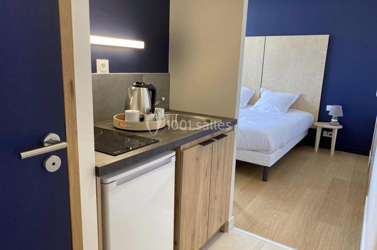 Kitchenette équipée avec plaque de cuisson, évier, bouilloire et vue sur une chambre avec lit double et mur bleu.