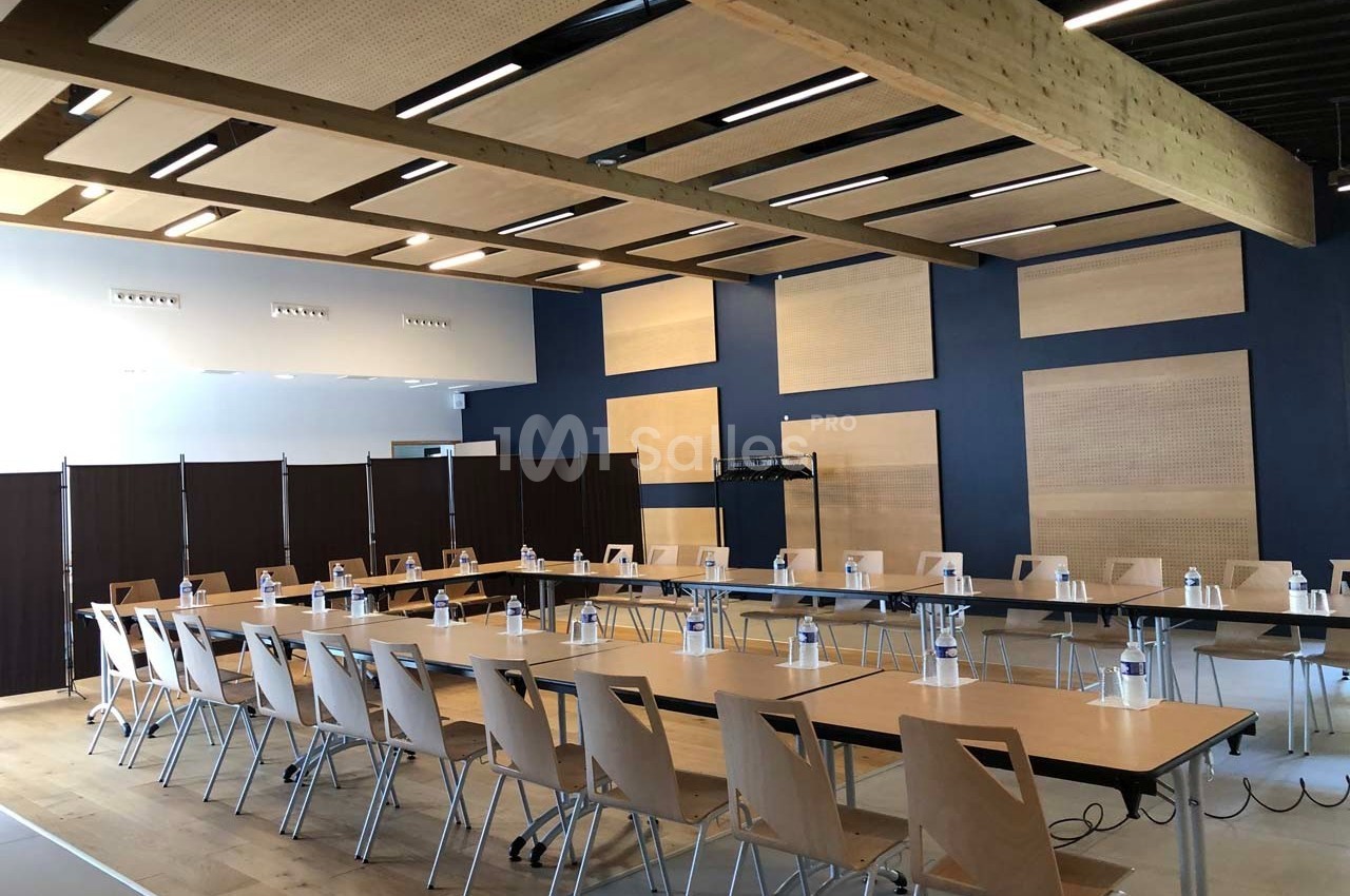 Salle de réunion lumineuse avec tables disposées en U, chaises en bois clair et bouteilles d'eau alignées.