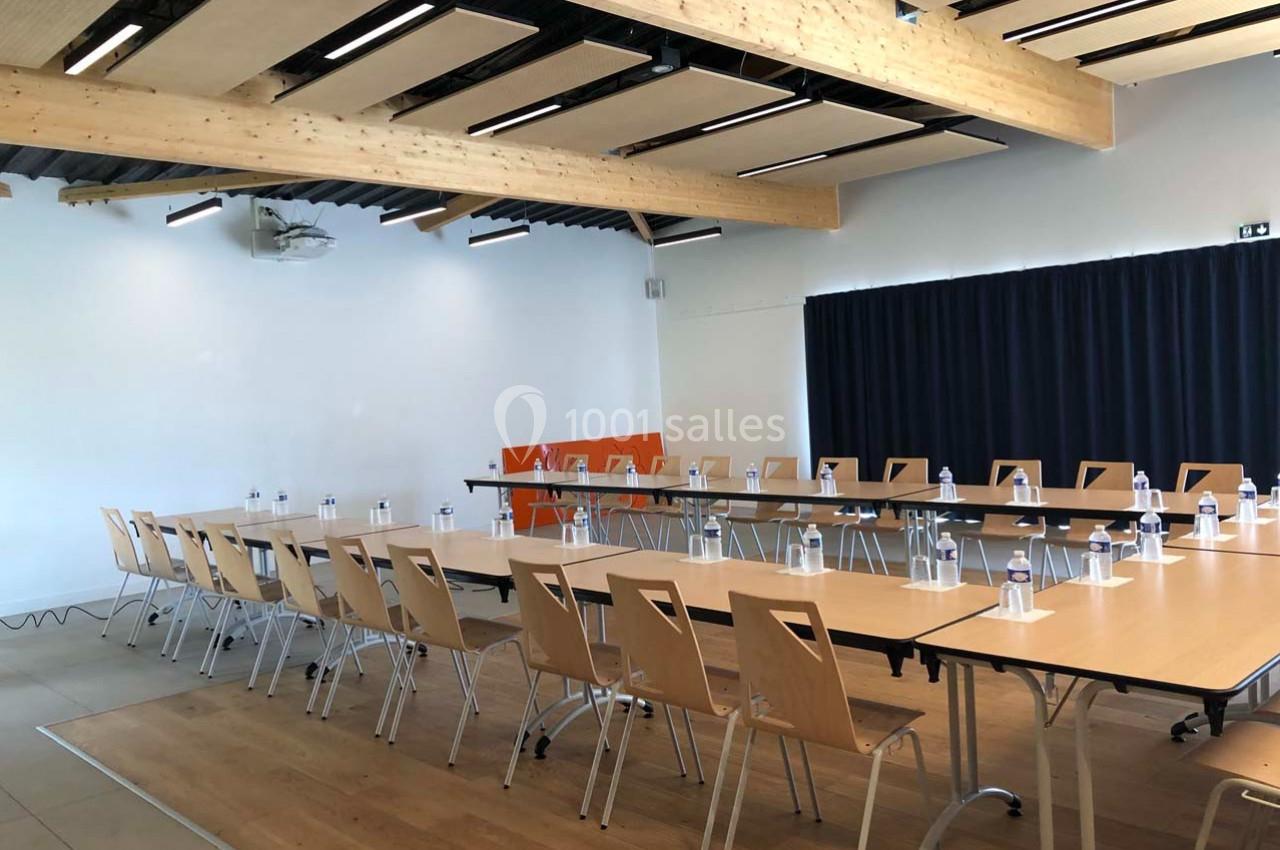 Salle de réunion lumineuse avec tables disposées en U, chaises en bois clair et bouteilles d'eau sur les tables.