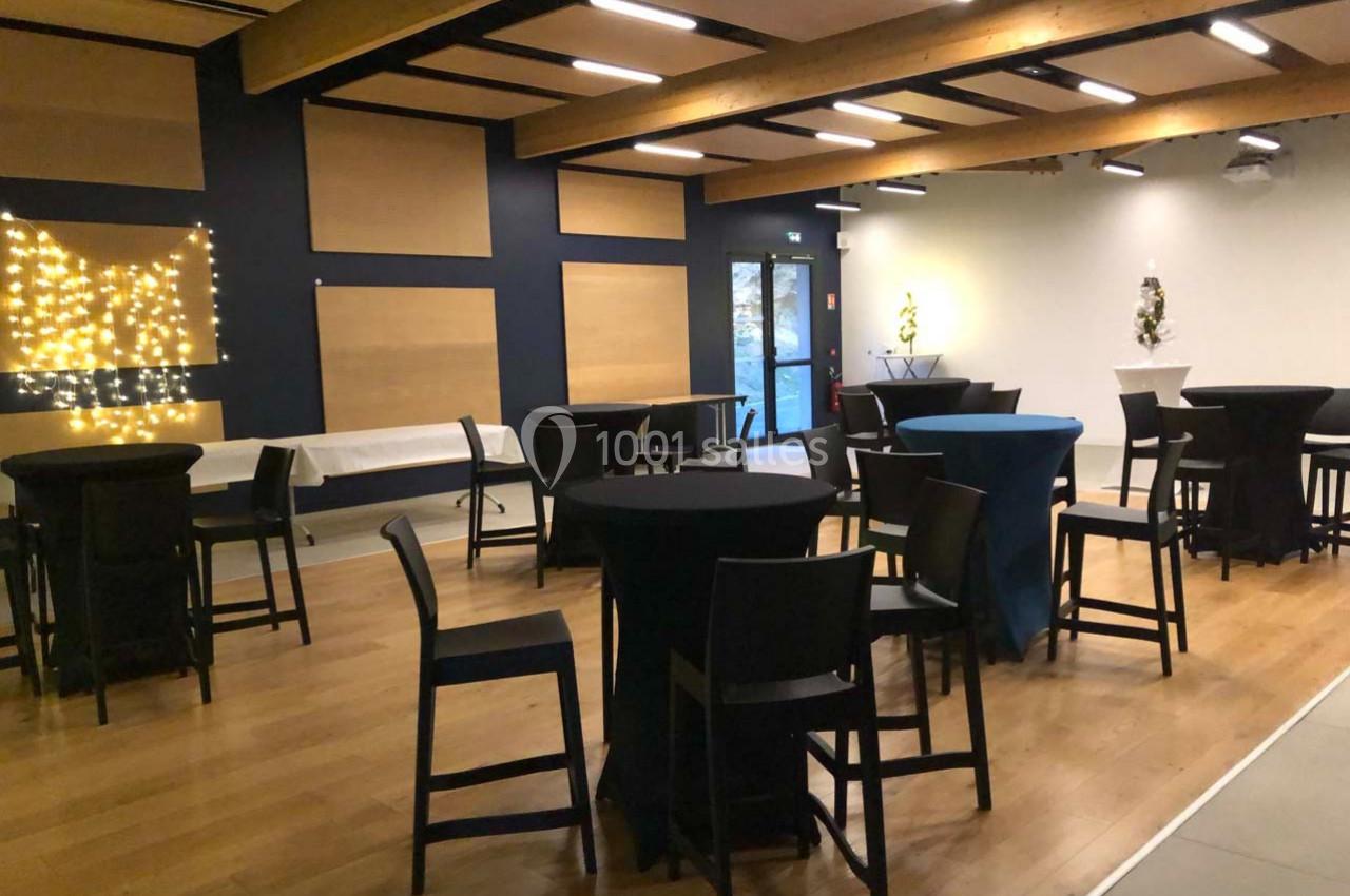 Salle aménagée avec des tables hautes et chaises noires, éclairée par des guirlandes lumineuses et des spots.