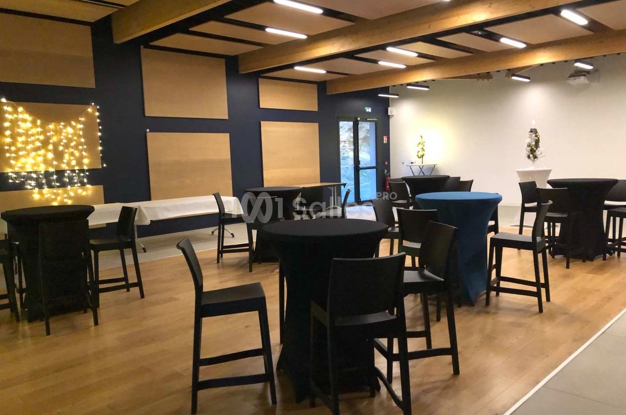 Salle aménagée avec des tables hautes et chaises noires, éclairée par des guirlandes lumineuses et des spots.