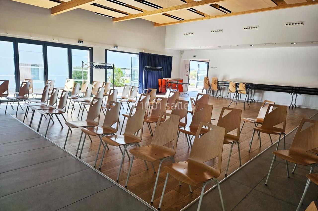 Salle lumineuse avec des rangées de chaises en bois vides, grandes baies vitrées et plafond avec poutres apparentes.