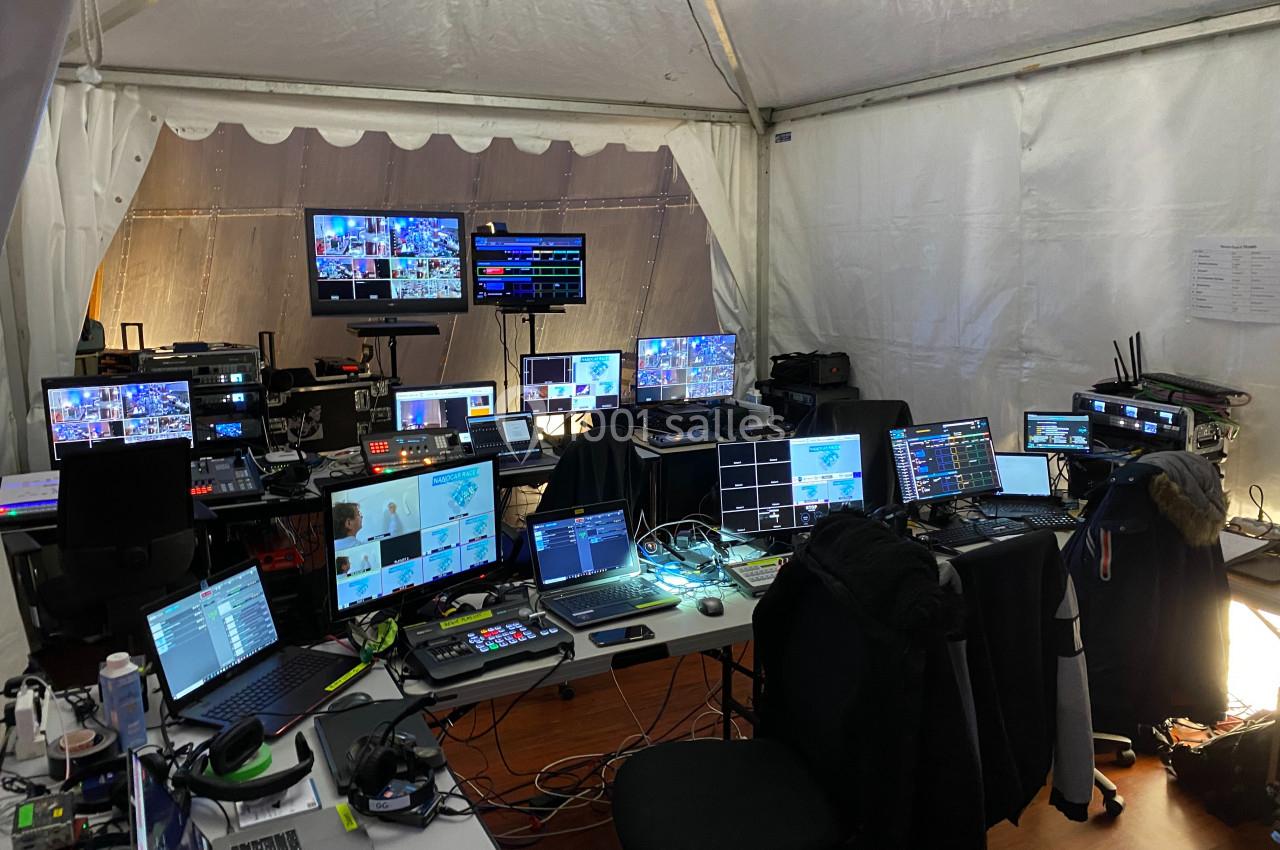 Poste de contrôle audiovisuel sous une tente, avec plusieurs écrans, ordinateurs et équipements techniques.