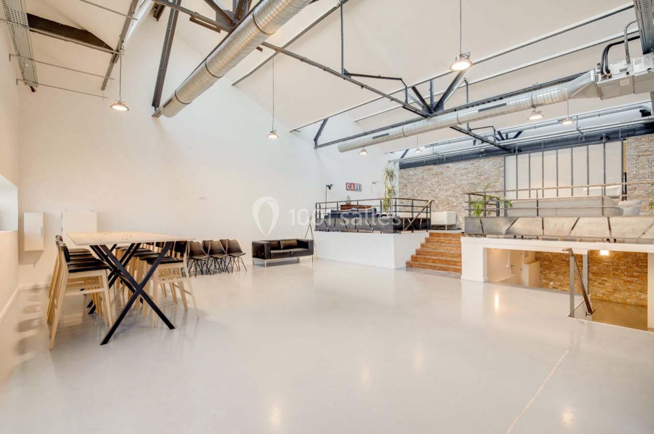 Espace intérieur lumineux avec sol blanc, murs en briques, mezzanine, tables, chaises et poutres apparentes.