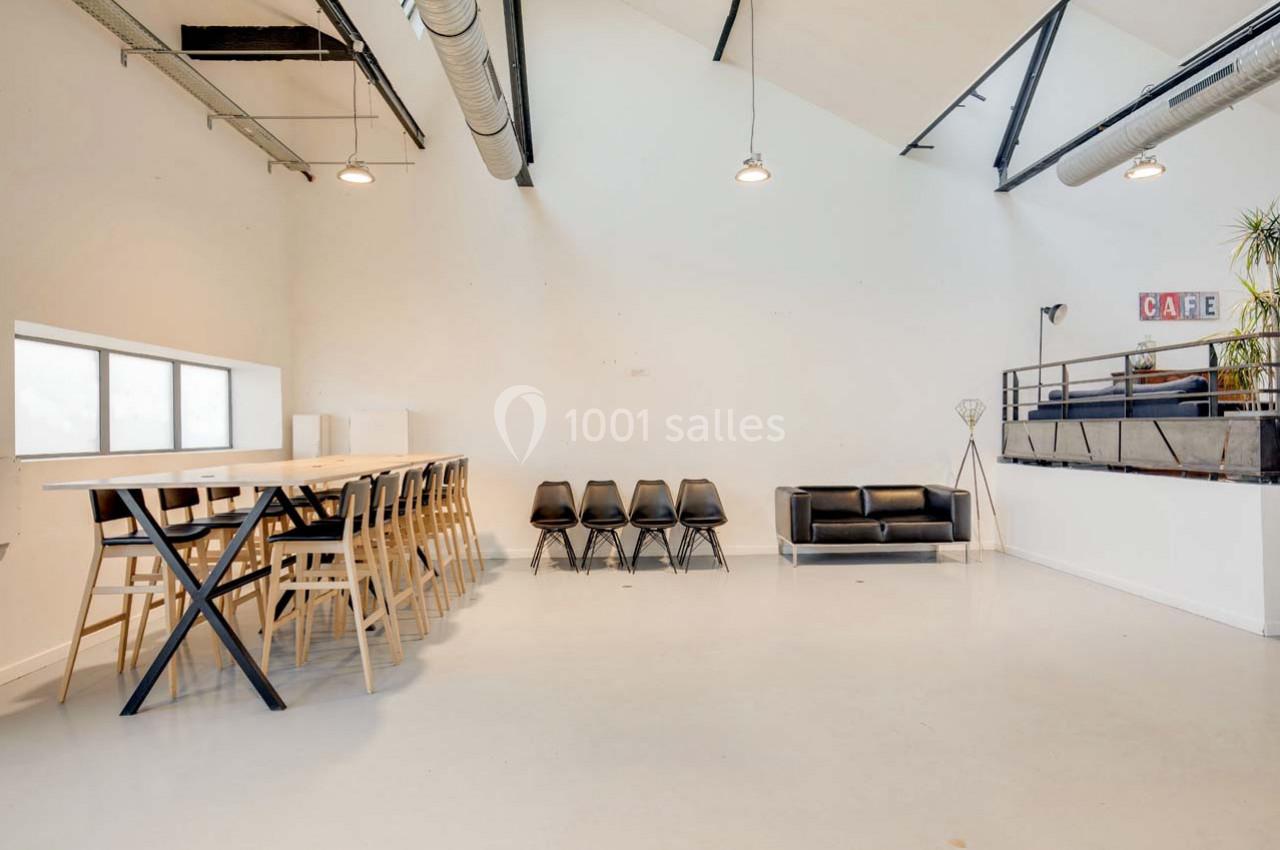 Salle lumineuse avec grande table, chaises alignées, canapé noir et mezzanine avec décoration minimaliste.
